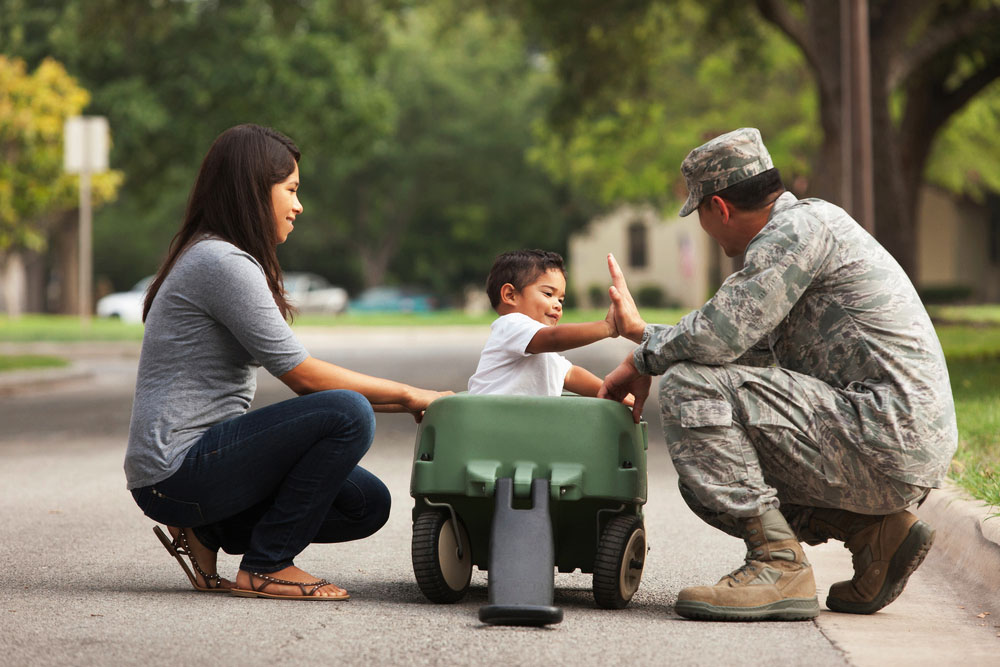 Male servicemember in uniform kneeling beside a wagon with a child inside. Female spouse in civilian clothes kneels on the opposite side of the wagon. 